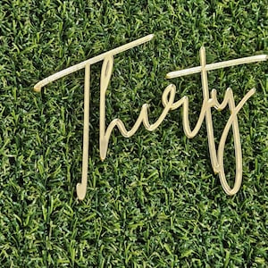 May include: A gold-coloured acrylic cake topper featuring the word Thirty written in a cursive script font. The letters are connected and have a reflective, smooth surface finish. The design is lightweight and features a thin, elongated structure.