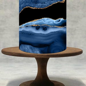 Puede incluir: Pantalla de lámpara cilíndrica con diseño de ágata azul y detalles dorados, exhibida sobre un soporte de pastel de madera. La pantalla presenta una combinación de colores azul oscuro y negro con vetas doradas, creando un contraste llamativo. El soporte es de madera oscura.