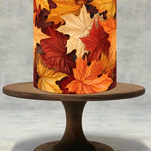 Pode incluir: Abajur cilíndrico com um vibrante design de folhas de outono em tons de vermelho, laranja, amarelo e creme. O abajur está sobre um suporte de bolo de madeira marrom escuro, criando uma estética quente e sazonal. O suporte tem uma parte superior e inferior redondas, conectadas por um pedestal curvo.