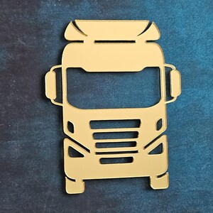 Può includere: Un ritaglio dorato e stilizzato di un autoarticolato. Il camion presenta un frontale dettagliato con fari, una griglia e una cabina. Il design è una rappresentazione piatta e bidimensionale di un veicolo commerciale.