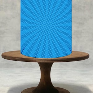 Könnte beinhalten: Ein zylindrisches blaues Objekt mit einem Comic-Design aus radialen Linien und Punkten, platziert auf einem runden Kuchenständer aus Holz. Der Ständer hat eine dunkelbraune Oberfläche und einen Sockel.
