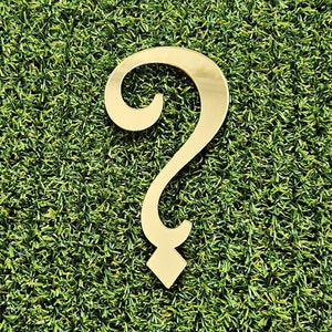 May include: A gold-coloured, stylised question mark, with a curved top and a diamond-shaped base, is set against a backdrop of green artificial turf. The reflective surface of the question mark creates a bright, eye-catching effect.