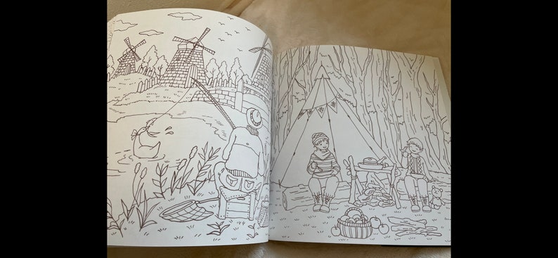 Puede incluir: Un libro para colorear abierto en dos p&aacute;ginas. Una p&aacute;gina representa a una persona pescando cerca de molinos de viento y un estanque con un pato. La otra p&aacute;gina muestra a dos personas acampando cerca de una tienda de campa&ntilde;a y una cesta de manzanas. Las ilustraciones son en blanco y negro.