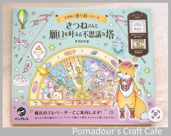 May include: A light blue coloring book titled きつねさんと願いを叶える不思議な塔 by rocca, featuring a central semi-circular illustration of a magical tower, a fox character in a red hat and cape, and various small icons like a hot air balloon, pudding, and keys.