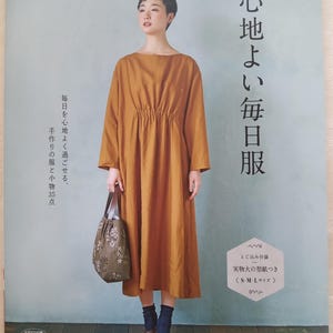 Pode incluir: Uma mulher em um vestido mostarda de mangas compridas, segurando uma sacola floral. A imagem é de um livro intitulado "Roupas confortáveis ​​para o dia a dia" com texto em japonês. A capa do livro também inclui texto sobre padrões e tamanhos.