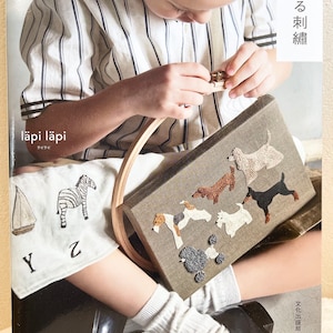 以下が含まれることがあります： 若い人が、針仕事の作品に取り組んでいます。作品は、刺繍された犬が様々な種類描かれた小さなキャンバスです。キャンバスは、木製の刺繍枠に入っています。その人は、ストライプのシャツと白い靴下を履いています。本の文字は「läpi läpi」と「夢みる 刺繍」です。