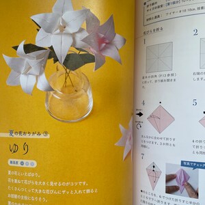 May include: A step-by-step guide on how to fold a white origami lily. The instructions include diagrams and text in Japanese. The finished origami lily is shown in a clear glass vase with green stems.