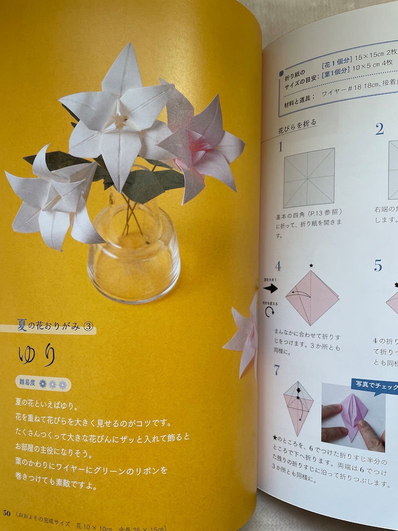 May include: A step-by-step guide on how to fold a white origami lily. The instructions include diagrams and text in Japanese. The finished origami lily is shown in a clear glass vase with green stems.