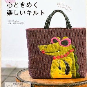 Könnte beinhalten: Ein Buchcover mit einer lila und weiß gepunkteten Tragetasche mit einer grünen Alligator-Applikation. Der Alligator trägt eine rosa Sonnenbrille und eine rote Perlenkette. Der Buchtitel lautet "キュンと! 心ときめく 楽しいキルト" und der Untertitel lautet "ここほれわんわん 丸濱淑子・由紀子". Das Buchcover enthält auch den Text "レディブティックシリーズ" und "実物大の型紙 アップリケの縫い方順序つき".