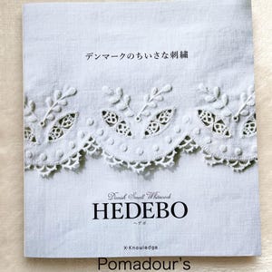 Può includere: Un libro bianco con il titolo "Danish Small Whitework HEDEBO" e il testo "デンマークの ちいさな 刺繍" in giapponese. La copertina del libro presenta un primo piano di un delicato ricamo bianco su un tessuto di lino bianco.