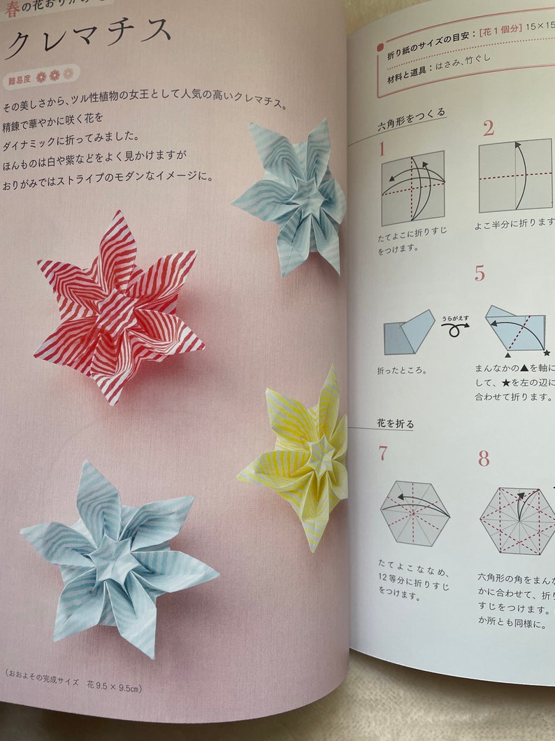 May include: Instructions for folding a paper origami flower in the shape of a clematis. The instructions are written in Japanese. The origami flower is shown in three different colors: red and white stripes, yellow and white stripes, and blue and white stripes. The finished origami flower is 9.5 inches by 9.5 inches.