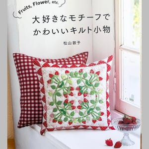 Puede incluir: Una portada de libro de manualidades con un cojín blanco con un patrón de acolchado de fresas rojas y verdes y un cojín de cuadros rojos y blancos detrás. El texto dice "Fruits, Flower, etc.", "大好きなモチーフでかわいいキルト小物" y "松山敦子".