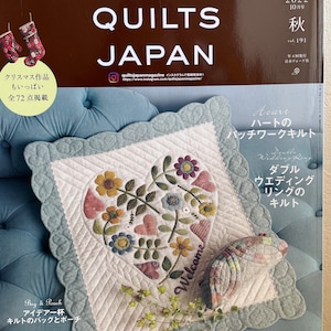Peut inclure: Une couverture de magazine pour Quilts Japan, une publication sur la courtepointe. La couverture présente une courtepointe en forme de cœur avec un motif floral et les mots "Bienvenue" brodés dessus. Le magazine est intitulé "Quilts Japan" et le numéro du magazine est 191. La couverture présente également une courtepointe plus petite en forme de poisson.