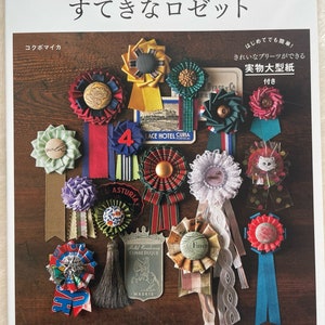 Puede incluir: Un libro titulado "Coser sin coser: Maravillosas rosetas" con una muestra colorida de rosetas hechas de tela y cinta. La portada del libro presenta el texto japonés "コクボマイカ" y "実物大型紙付き", que se traduce como "Patrón de tamaño real incluido".