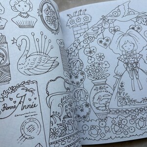 May include: An open book with detailed black and white illustrations. The drawings include a swan pin cushion, a spool of thread with a horse emblem, a doll, floral designs, and the text "Bonne Ann&eacute;e". The style is reminiscent of vintage coloring books.