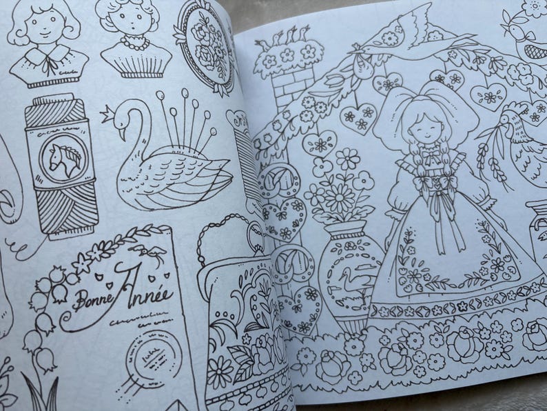 May include: An open book with detailed black and white illustrations. The drawings include a swan pin cushion, a spool of thread with a horse emblem, a doll, floral designs, and the text "Bonne Ann&eacute;e". The style is reminiscent of vintage coloring books.