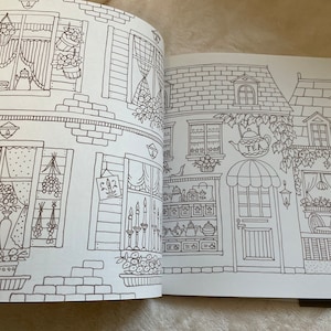 Puede incluir: Un libro para colorear abierto en una doble p&aacute;gina con dibujos lineales detallados de edificios. Las ilustraciones incluyen ventanas, puertas, arreglos florales y un letrero que dice "TEA". Se ve un gato en una ventana.