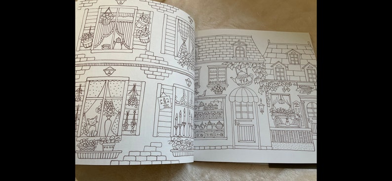 Puede incluir: Un libro para colorear abierto en una doble p&aacute;gina con dibujos lineales detallados de edificios. Las ilustraciones incluyen ventanas, puertas, arreglos florales y un letrero que dice "TEA". Se ve un gato en una ventana.