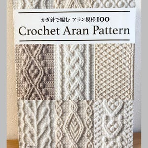 以下が含まれることがあります： 「Crochet Aran Pattern」というタイトルの本で、日本語のテキストが書かれています。表紙には、クリーム色とベージュ色の様々なテクスチャードかぎ針編みのステッチパターンが表示されています。Pomadour's Craft Cafeからの本です。