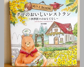 May include: A colorful illustrated book cover featuring a bear chef holding a salad bowl. The title reads "Kuma no Oishii Restaurant" with Japanese text and English text "GINO the bear, a wonderful chef." The cover includes a house, garden, and yellow flowers.