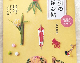 May include: A book cover featuring various handcrafted Japanese Mizuhiki art pieces in vibrant colors. The cover includes text in Japanese and showcases decorative knots, floral designs, and other ornamental creations. The background is a light yellow.