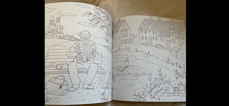 Puede incluir: Una doble p&aacute;gina de un libro para colorear con dibujos lineales detallados. Una p&aacute;gina muestra a un hombre tocando el acorde&oacute;n en un banco del parque con un gato y una bolsa. La otra p&aacute;gina representa casas, un lago y un picnic.