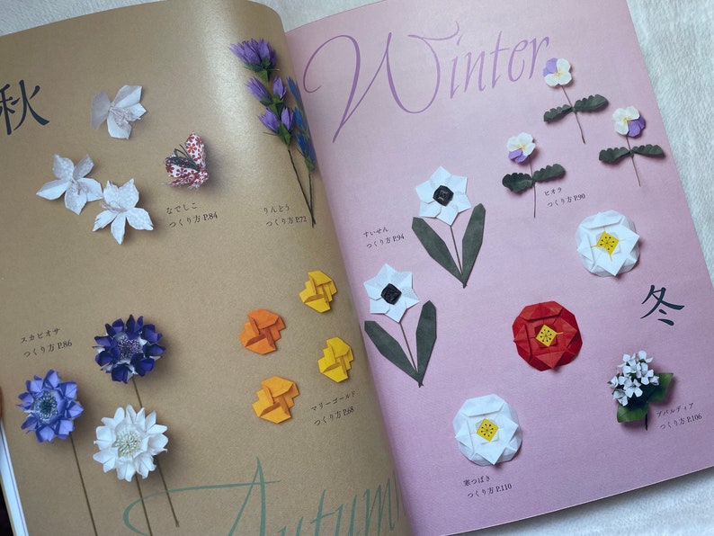 May include: A book with origami flower instructions. The book has a pink background with the word "Winter" written in black. The book also has a brown background with the word "Autumn" written in black. The book features origami flowers in various colors, including white, red, purple, and yellow.