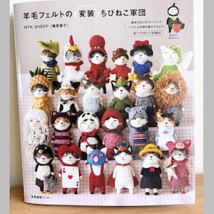 Può includere: Una copertina di libro con una collezione di figurine di gatti in feltro fatte a mano vestite con vari costumi. I gatti sono disposti in file, mostrando una varietà di abiti, inclusi costumi a tema animale e personaggio. Il titolo del libro è in giapponese.