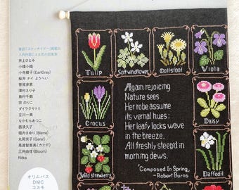 Op de afbeelding: Een kruissteekpatroonboek met bloemmotieven, waaronder tulpen, viooltjes en madeliefjes. De omslag toont een zwarte banner met geborduurde bloemenillustraties en tekst in het Japans en Engels. Uitgegeven door een Japans bedrijf.