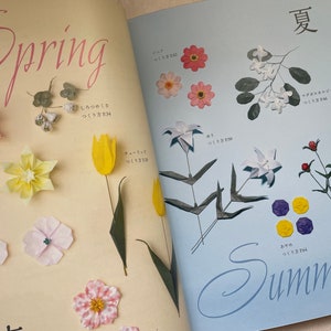 May include: A book with origami flower designs. The left page shows spring flowers, including a red origami star, a yellow origami tulip, and a pink origami flower. The right page shows summer flowers, including a white origami jasmine, a white origami lily, and a purple origami flower.