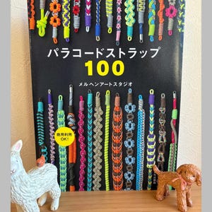 Op de afbeelding: Een boek getiteld "Paracord Straps 100" met verschillende kleurrijke paracord armband ontwerpen. De omslag is zwart met Japanse tekst en het nummer 100 in geel. Decoratieve beeldjes van een lama en een hond staan op de voorgrond.