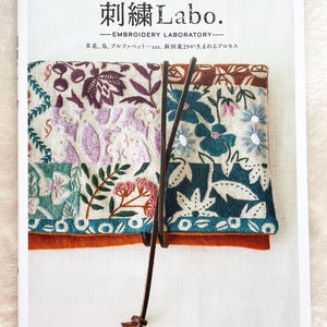 以下が含まれることがあります： 「刺繍Labo」と書かれた日本語の本。表紙には、水色、紫、白の花柄の刺繍が施されています。茶色の革紐で本が留められています。白い背景に本が置かれています。本の表紙には花柄のデザインがあります。