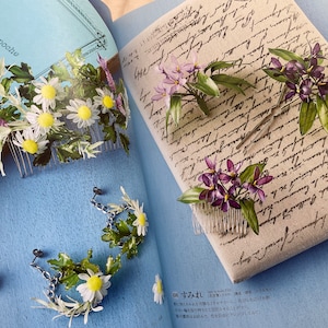 May include: Two hair combs with floral designs. One comb features white daisies and green leaves, the other features purple flowers and green leaves. The combs are on a blue background.