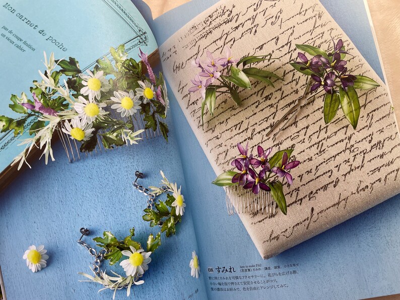 May include: Two hair combs with floral designs. One comb features white daisies and green leaves, the other features purple flowers and green leaves. The combs are on a blue background.