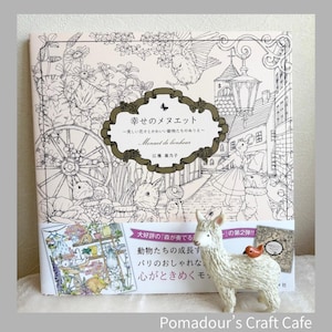 May include: A coloring book titled "Menuet de bonheur" with Japanese text and illustrations of flowers and animals. A small white llama figurine with a red bird on its back is in front of the book. The book cover has a floral design.