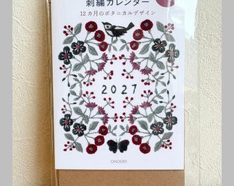 May include: A 2027 spiral-bound calendar featuring embroidered botanical designs with red flowers, gray leaves, a black bird, and a black butterfly. The cover text includes Alice Makabe, 刺繍カレンダー, 12カ月のボタニカルデザイン, and ONDORI. The calendar is encased in a clear plastic sleeve.