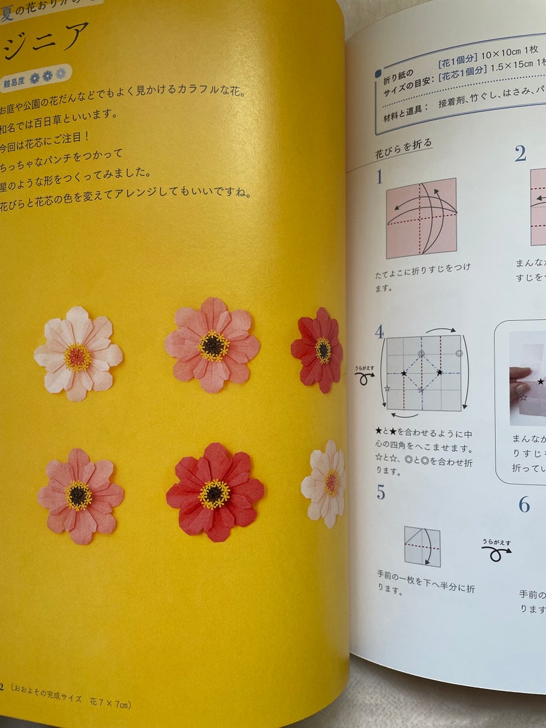 May include: A step-by-step guide on how to fold a paper zinnia flower. The instructions are written in Japanese and include diagrams showing the folding process. The finished flower is pink with a brown center.