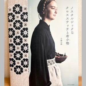May include: A book titled "CROSS STITCH VOYAGE" with a cover featuring a woman and Japanese text. The spine has a beige fabric with a black and white floral cross-stitch design. The book is on a wooden surface.