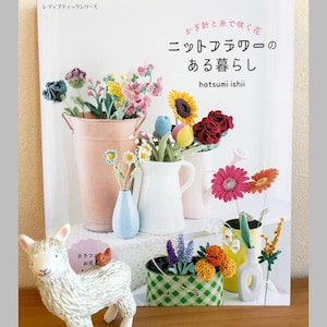 以下が含まれることがあります： 「ニットフラワーのある暮らし」というタイトルの本。日本語のテキストと、花瓶や容器に入ったかぎ針編みの花。白いセラミックのラマの置物と、背中に小さなオレンジ色の動物がいます。