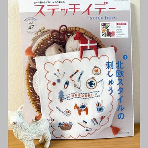 May include: A special issue of "Stitch Idées" magazine with a white embroidered textile featuring various embroidery designs, a red wooden horse, and a small white ceramic sheep figurine. The magazine is vol. 47.