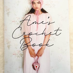 Può includere: Un libro intitolato "Ame's Crochet Book" con uno sfondo floreale rosa. La copertina presenta una giovane persona che indossa un cappello e un vestito all'uncinetto rosa, tenendo una borsa all'uncinetto.