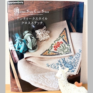 Puede incluir: Un libro titulado "Antique Style Cross Stitch" con texto japonés, que presenta patrones de punto de cruz. La imagen muestra una pieza parcialmente cosida con diseños florales y de conejos, junto con carretes de hilo y una figura decorativa de llama.