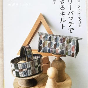 May include: A book cover with the title "Three Patch Quilts" in Japanese. The cover features a wooden basket, a wooden doll, and a fabric pouch, all made with patchwork quilting techniques.