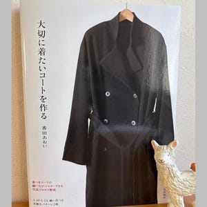Puede incluir: Portada de libro con un diseño de abrigo negro colgado de una percha de madera. El título del libro está en japonés, con texto adicional. Una pequeña figura decorativa de llama está sobre una superficie de madera. El libro trata sobre la costura de abrigos.