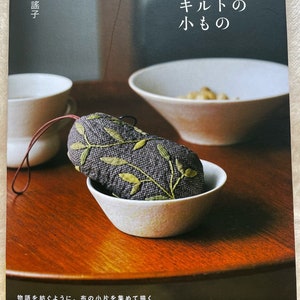 May include: A book titled "語りかけるキルトの小もの" by 斉藤謠子. The book cover features a close-up of a small, quilted, fabric pouch with green embroidery on a dark grey background. The pouch is sitting in a white ceramic bowl on a wooden table.