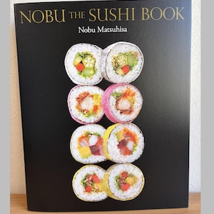 Può includere: Un libro di cucina intitolato "NOBU THE SUSHI BOOK" di Nobu Matsuhisa. La copertina è nera con scritte dorate e presenta colorati rotoli di sushi. I rotoli di sushi sono disposti in file, mostrando vari ingredienti e presentazioni.