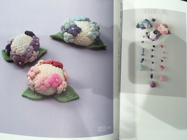 May include: Three felt flower embellishments in shades of pink, purple, and blue. The flowers are arranged on a light purple background. The flowers are made of felt and have a textured surface. The flowers are surrounded by green felt leaves.