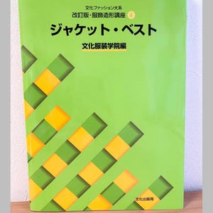 May include: A green book with Japanese text, featuring the title "ジャケット・ベスト" (Jacket & Vest) in large black characters. The cover has a pattern of yellow and green rectangles. The book is on a wooden surface.