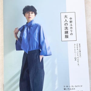 Op de afbeelding: Een persoon in een blauwe blouse en donkere broek poseert met een boek. De boekomslag heeft Japanse tekst en de titel "COUTURIER SEWING CLASS / YUKARI NAKANO". Het boek gaat over het naaien van kleding, waaronder blouses en broeken.