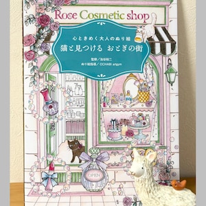 Könnte beinhalten: Ein Malbuch mit dem Titel "Rose Cosmetic Shop" mit japanischem Text und Illustrationen eines Kosmetikgeschäfts, Katzen und Schönheitsprodukten. Das Cover ist mit Rosen und einer dekorativen Parfümflasche verziert. Eine weiße Lama-Figur befindet sich im Vordergrund.